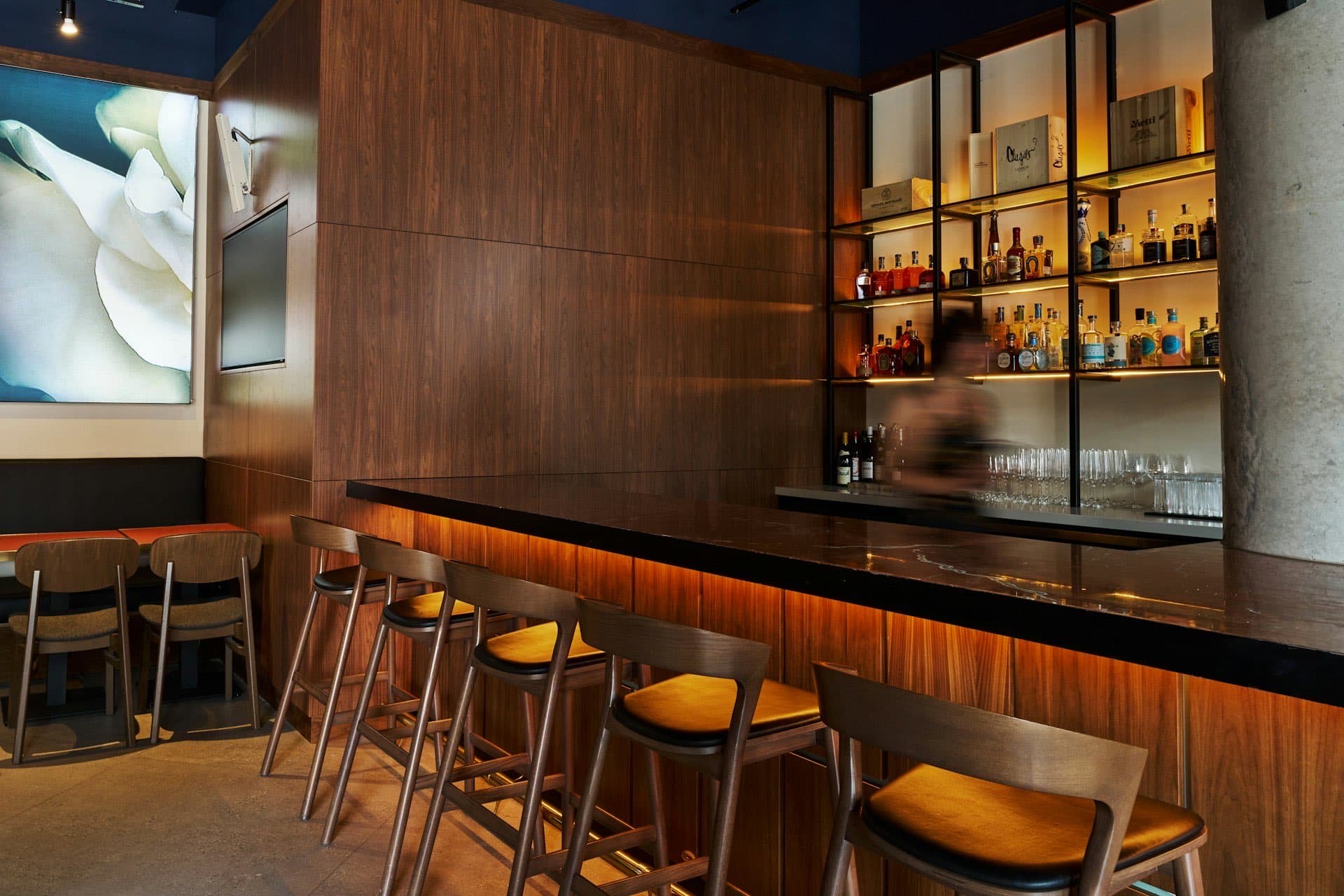 Upscale bar interior with a wood-paneled counter, stools, and backlit spirit shelves in warm amber light