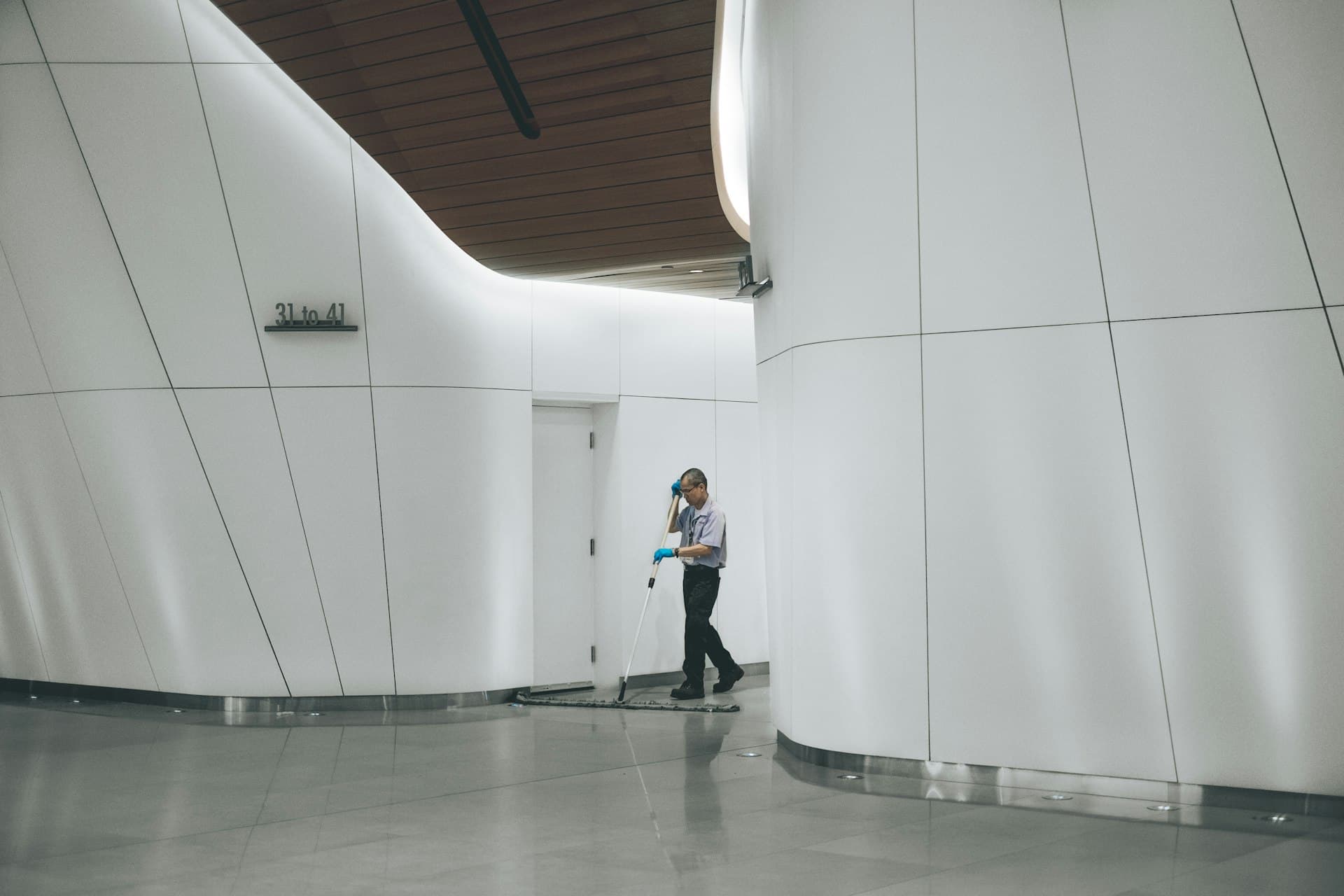 Janitorial worker in uniform mopping the polished floor of a large commercial building corridor with a flat mop