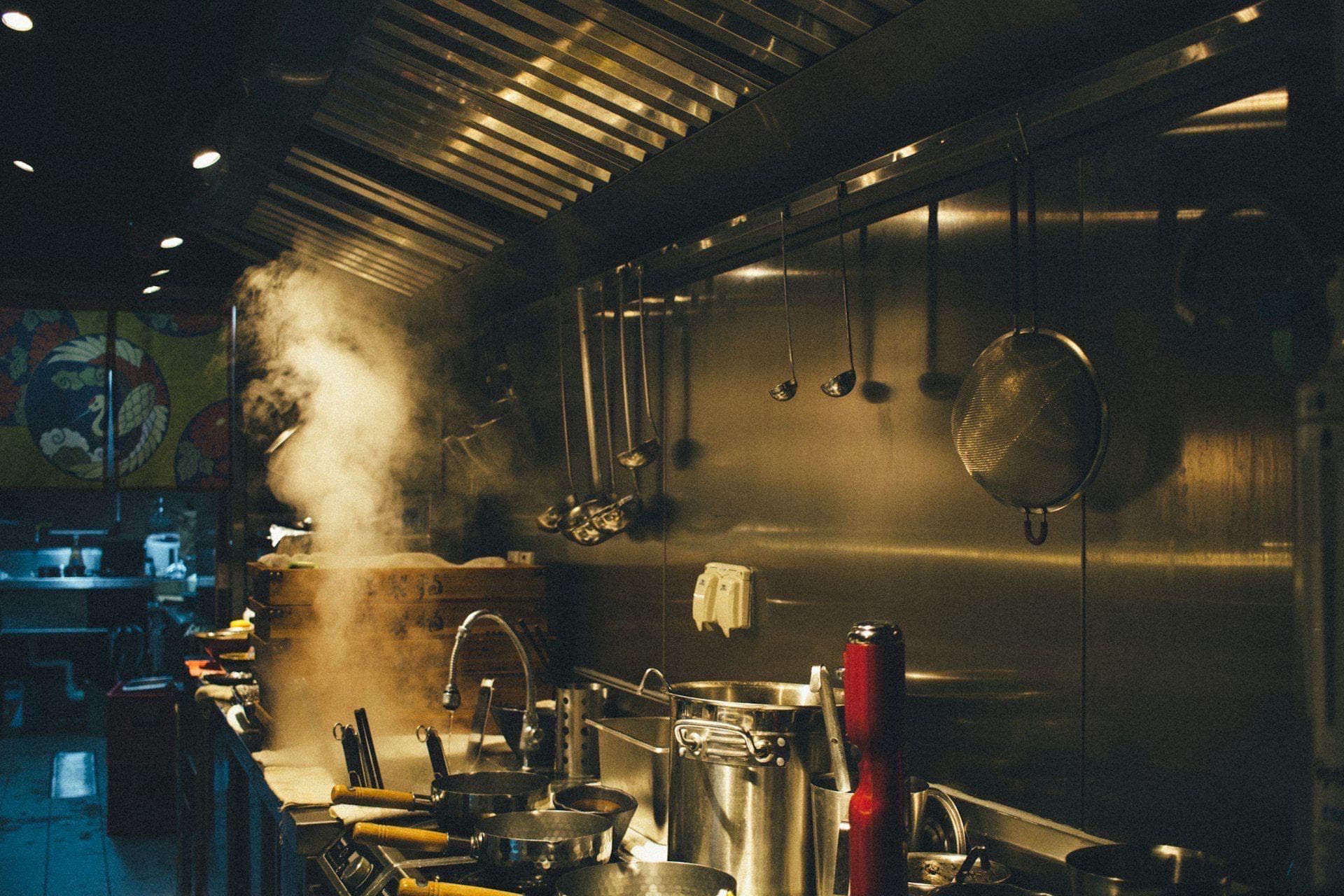 Commercial kitchen line mid-service with stainless cookware and hanging ladles, steam rising above the range