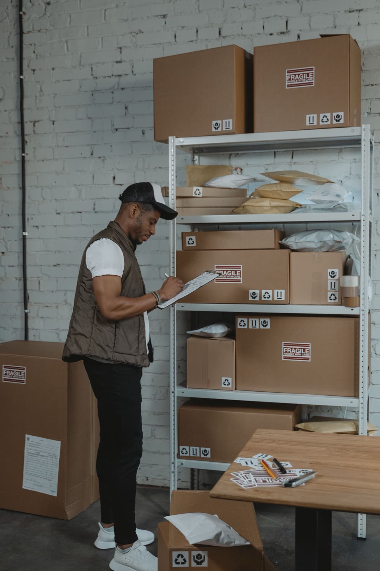 Delivery contractor in a vest checking a clipboard beside shelving stacked with labeled cardboard parcels at a distribution warehouse
