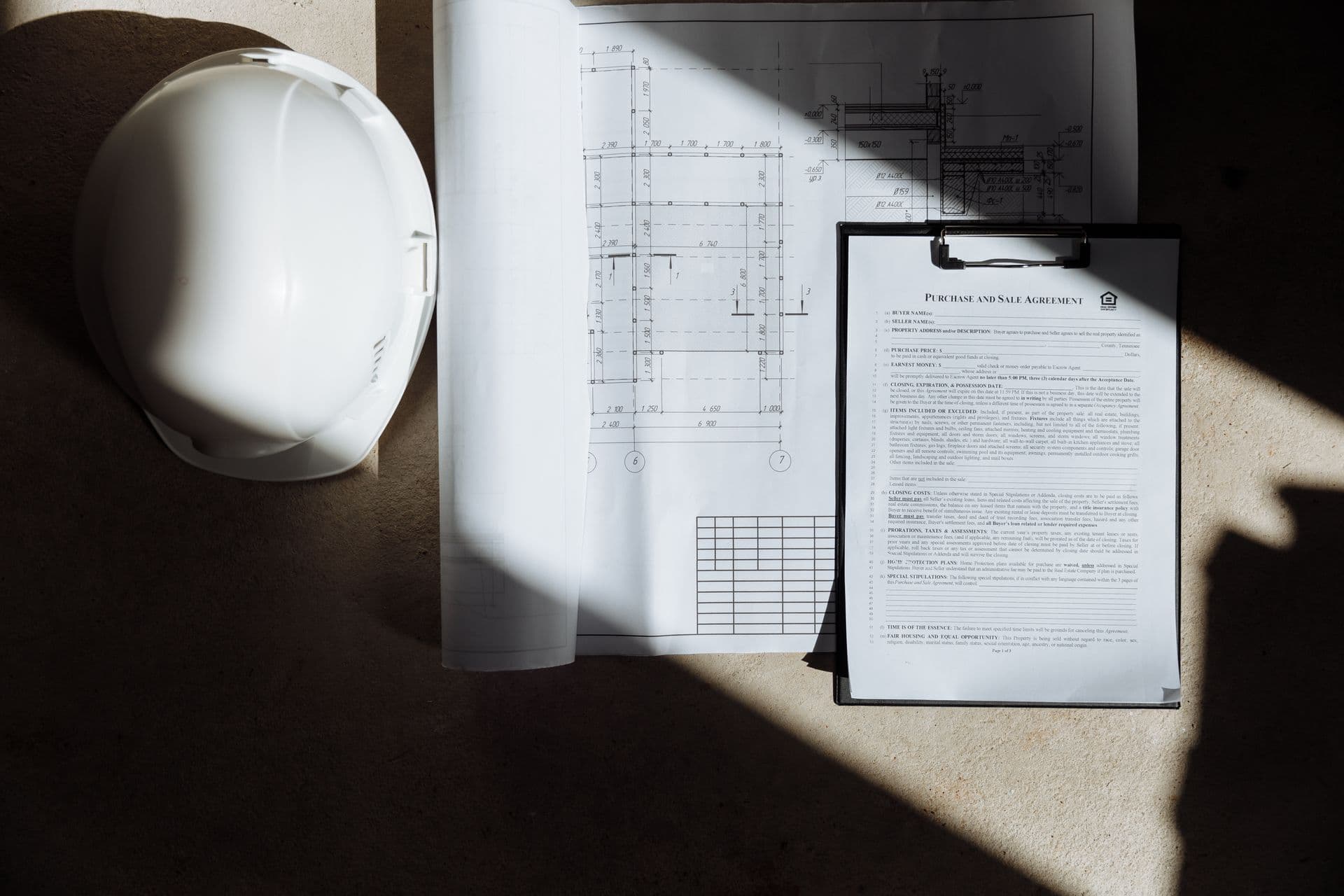 A white construction hard hat resting beside a rolled floor-plan blueprint and a clipboard on a jobsite table in raking light