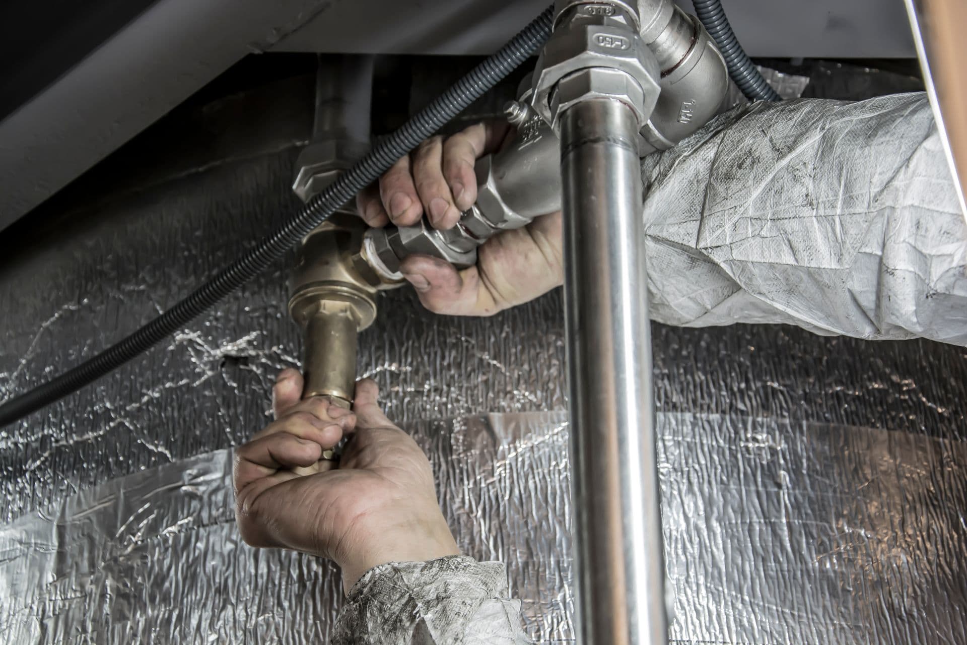 Plumber's hands installing and tightening brass and steel pipe fittings on an insulated commercial pipe run indoors