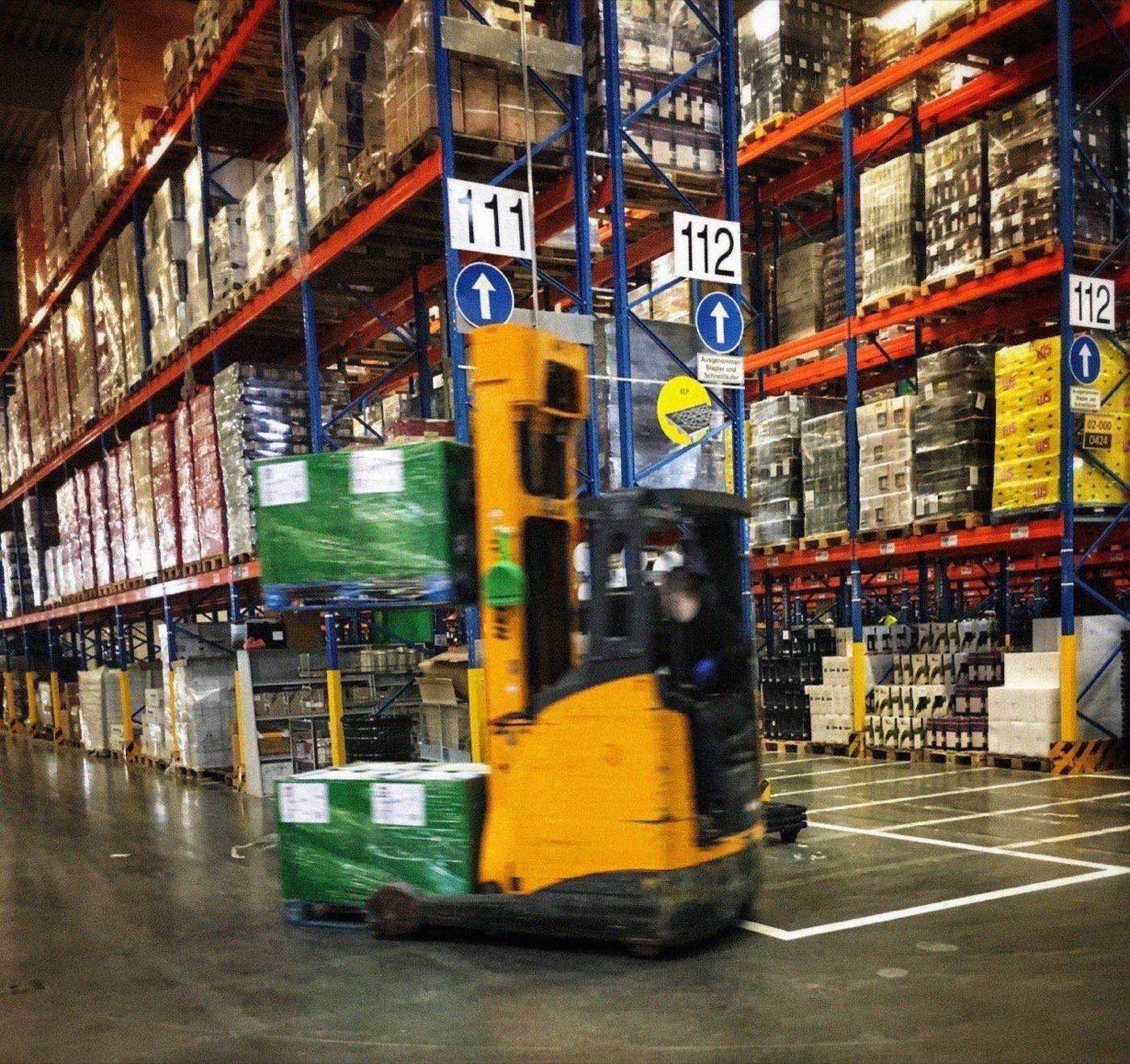 Reach-forklift operator moving a pallet through numbered aisles of a large warehouse with tall pallet racking